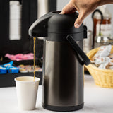 A hand presses down on the black lid of a stainless steel airpot, dispensing hot coffee into a white paper cup. In the background, vacuum insulated coffee supplies like sugar packets, creamers, and a basket with lids can be seen as steam rises from the cup.