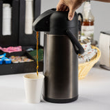 A hand presses the lid of a vacuum insulated stainless steel airpot dispenser, pouring coffee into a white paper cup. In the background are assorted coffee supplies, sweeteners, stirrers, cups, and a wicker basket with creamers and tea bags.