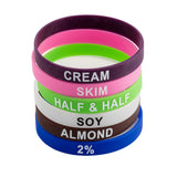 A stack of colorful TableCraft Products Silicone Flavor Bands, each labeled with a type of milk—purple CREAM, pink SKIM, green HALF & HALF, white SOY, brown ALMOND, and blue 2%—all with bold white text. Perfect for Cash & Carry displays.