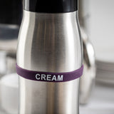 A stainless steel canister with a purple Silicone Flavor Band labeled CREAM in white letters, set against a blurred background with dishes and other kitchen items from TableCraft Products.