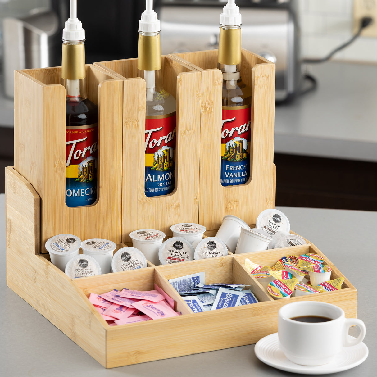 A coated bamboo coffee condiment organizer holds three Torani syrup bottles, assorted creamers, sugar packets, and sweeteners. A white cup of coffee rests nearby on a stainless steel countertop in a modern kitchen setting.