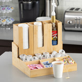 A coated bamboo coffee condiment organizer holds stacked paper cups, sugar packets, creamers, a syrup bottle with a pump, and coffee pods. A cup of tea sits in front, with kitchen appliances and cup lid holders visible in the background.