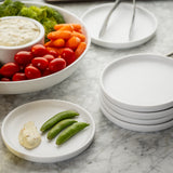 A small white melamine serving plate with a scoop of dip and two snap peas sits on a marble surface beside a stack of empty plates. In the background, the Terra Collection bowl holds broccoli, cherry tomatoes, carrots, and more dip. Metal tongs rest on the plates.