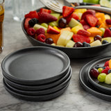 A stack of black plates sits on a marble countertop next to a large bowl of assorted fresh fruit, including strawberries, grapes, melons, and berries. A Terra Collection Serving Plate and serving utensils are visible with a glass of beverage in the background.