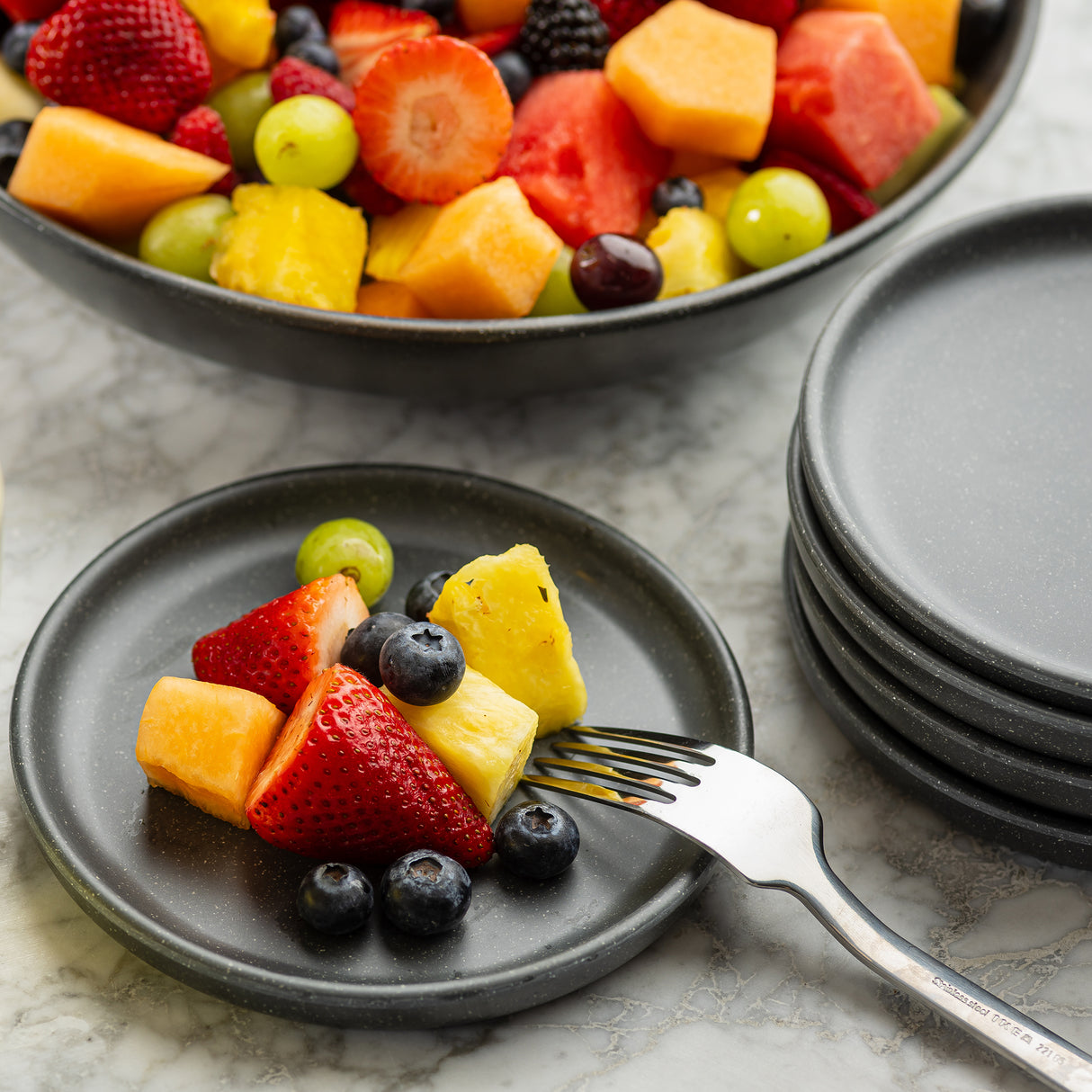 A small black melamine serving plate holds cut fruit—strawberry, pineapple, cantaloupe, grape, and blueberries. A fork rests on the plate. In the background sits a bowl of mixed fruit and stacked black plates atop a marble surface.