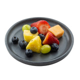 A dark gray speckled plate with assorted fresh fruit—blueberries, green grapes, strawberry halves, pineapple chunks, and a cantaloupe cube—arranged neatly on a white background.