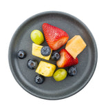 A gray speckled Terra Collection Serving Plate holds a small assortment of fruit: two strawberries, green grapes, blueberries, pineapple cubes, and cantaloupe cube, all viewed from above and isolated on a white background.