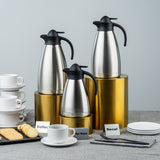 Three stainless steel carafes labeled Coffee, Hot Water, and Decaf sit on a gold nesting riser set. Nearby are stacked white cups, saucers, plates with cookies, napkins, and utensils, all on a gray tablecloth against a light gray background.