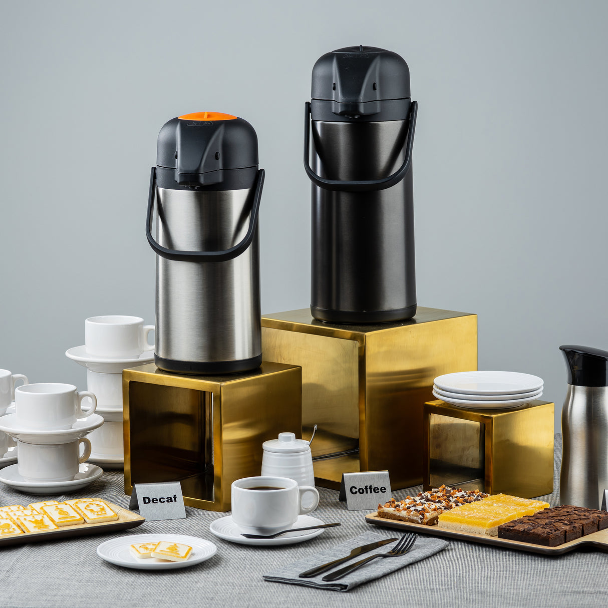 Two airpot coffee dispensers labeled “Decaf” and “Coffee” stand on a gold riser set among cups, plates, desserts, and utensils. Assorted pastries, stacked white dishes, and a pitcher complete the setup against a plain gray background.
