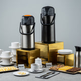 Two airpot coffee dispensers labeled “Decaf” and “Coffee” stand on a gold riser set among cups, plates, desserts, and utensils. Assorted pastries, stacked white dishes, and a pitcher complete the setup against a plain gray background.
