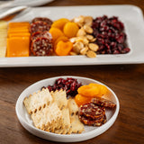 A small white melamine serving plate holds seeded crackers, dried sausage, apricots, and cranberries. Behind it, a larger platter from the Ridge Collection Serving Plate displays similar treats with cheddar and almonds on a wooden table.