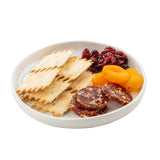 A white Ridge Collection Serving Plate from TableCraft Products displays rectangular seeded crackers, slices of dried sausage, dried apricots, and a small pile of dried cranberries, all neatly arranged in separate sections on a plain white background.