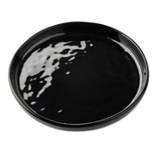 A round, glossy black ceramic serving plate with slightly raised edges sits on a white background. Light reflections highlight its smooth, shiny surface, emphasizing its minimalist and modern design. The plate is empty.