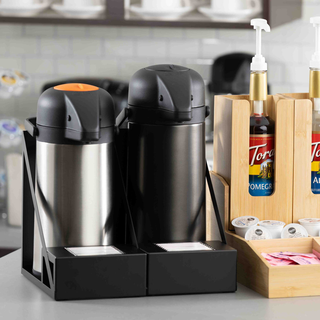 Two stainless steel airpots, one with an orange lever and one black, sit in powder coated steel holders with a stainless steel drip tray. Wooden racks hold flavored syrup bottles, creamer cups, and sweeteners. Shelves and cups line the background.