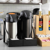 Two stainless steel airpots, one with an orange lever and one black, sit in powder coated steel holders with a stainless steel drip tray. Wooden racks hold flavored syrup bottles, creamer cups, and sweeteners. Shelves and cups line the background.