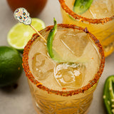 A cocktail in a patterned glass with a chili powder rim, ice cubes, and a jalapeño slice garnish. A Day of the Dead skull decorative pick adds flair. A lime wheel and whole limes are nearby; another similar drink is partially visible.