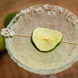 A close-up of a margarita in a glass with a salted rim, topped with a lime wheel on a bamboo pick decorated with a colorful Day of the Dead sugar skull. The drink appears icy and frozen, while the background is softly blurred.