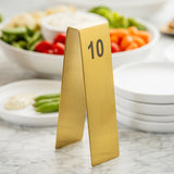 A gold Number Tent with the number 10 in black sits on a marble surface. Behind the stylish sign, stacked white plates and a bowl of assorted vegetables like tomatoes, green beans, and broccoli complete the scene.