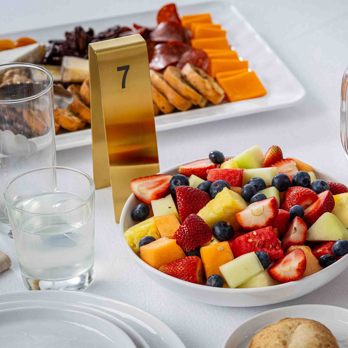 A white bowl filled with assorted fresh fruit sits on a white table next to a glass of water, a bread roll, and a gold TableCraft Number Tent 7. In the background is a cheese and meat platter.