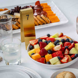 A white bowl filled with assorted fresh fruit sits on a white table next to a glass of water, a bread roll, and a gold TableCraft Number Tent 7. In the background is a cheese and meat platter.