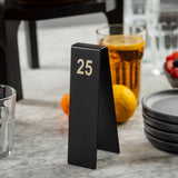 A black, triangular number tent from TableCraft Products, made of powder coated stainless steel with the number 25 in white, sits on a marble table beside a glass of amber liquid, stacked plates, fruits, and a small bowl of berries.