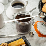 A glass jar filled with ground coffee sits open on a marble surface, with a long handle stainless steel coffee scoop resting on top. A white cup of black coffee, pastries in a black basket, and a plate of sweets complete the inviting breakfast scene.