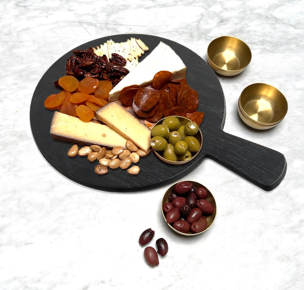 A black round serving paddle from the Onyx Melamine Collection displays crackers, dried apricots, cheese, spiced nuts, green olives, brown salami slices, and pecans. Three small brass bowls hold more olives beside the melamine board on a white marble surface.