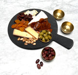 A black round serving paddle from the Onyx Melamine Collection displays crackers, dried apricots, cheese, spiced nuts, green olives, brown salami slices, and pecans. Three small brass bowls hold more olives beside the melamine board on a white marble surface.
