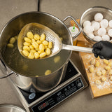 A hand in a black glove uses a TableCraft 12830 scoop colander to lift cooked gnocchi from boiling water on an induction cooktop. Nearby, eggs, an oil bottle, and floured gnocchi pieces rest on a wooden board.