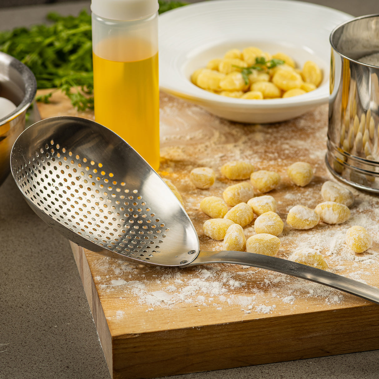 A TableCraft 12830 scoop colander rests on a floured wooden board with uncooked gnocchi. A bowl of cooked gnocchi, oil, a sifter, eggs, and fresh herbs set the scene for delicious homemade cooking.