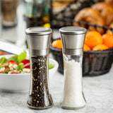 Two glass salt and pepper mills with metal tops from TableCraft Products sit on a marble counter; one holds black peppercorns, the other coarse salt. In the blurred background are a salad bowl and a black basket of oranges and bread rolls.