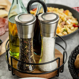 Two glass salt and pepper mills from TableCraft Products, topped with stainless steel, rest in a round black wire holder. In the background, a dish of pasta, oil bottle, and bread are arranged on a marble surface.