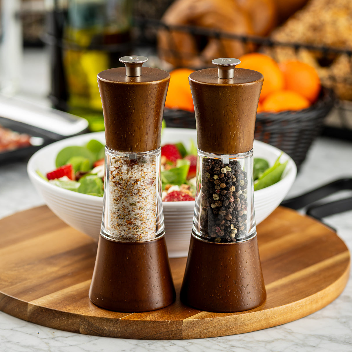 Two wooden Salt & Pepper Mill Set grinders, crafted with an acacia finish, stand on a cutting board before a salad bowl. The left holds coarse salt; the right, black peppercorns. Oranges in a basket and kitchen items are visible in the background.