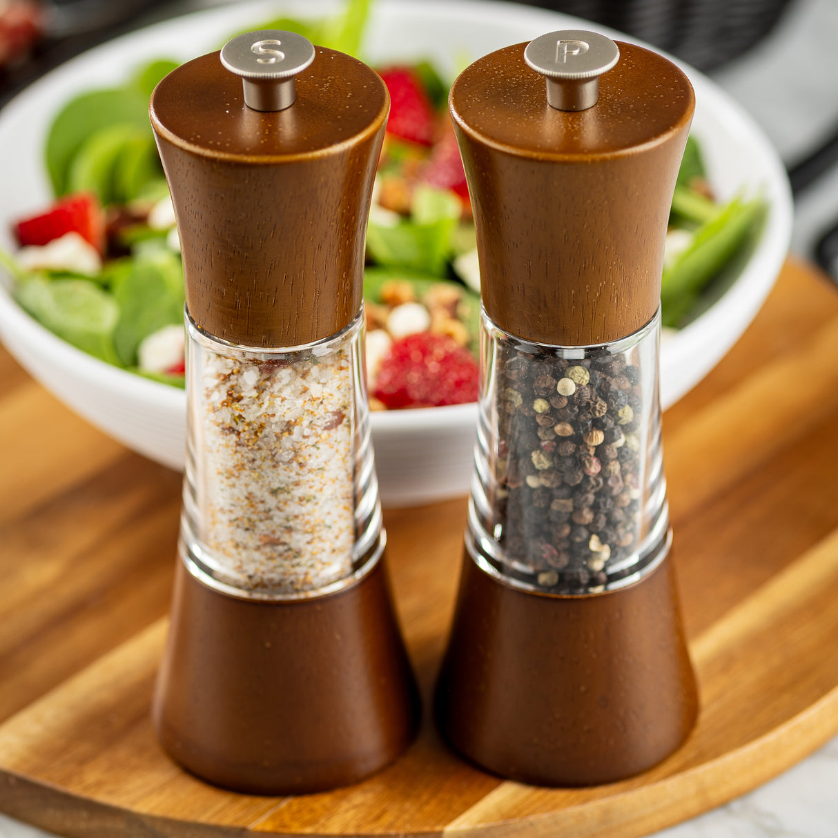 Two wooden salt and pepper grinders with a sleek acacia finish sit side by side on a wooden board. This Salt & Pepper Mill Set is crafted from durable rubberwood, with coarse salt on the left and whole black peppercorns on the right.
