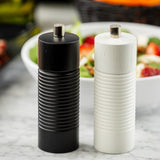 Two ribbed cylindrical grinders—one black, one white—from the TableCraft Products Salt & Pepper Mill Set stand side by side on a marble countertop. In the background, a white bowl brims with a colorful salad of greens and strawberries.