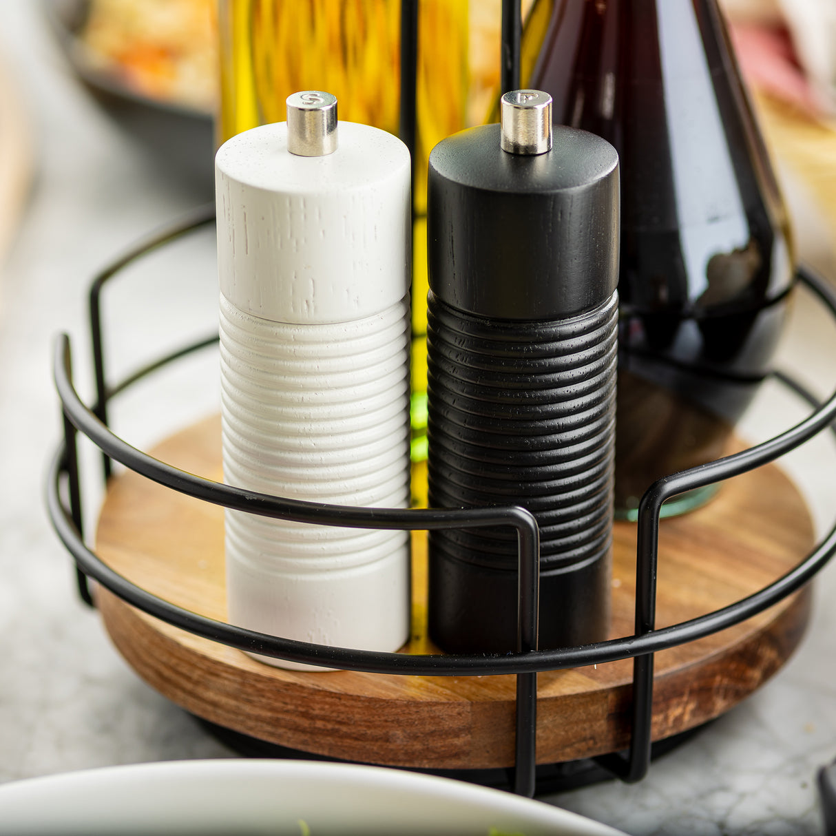 A close-up of a TableCraft Products Salt & Pepper Mill Set with white and black wood salt and pepper grinders in a metal and wooden holder, plus glass bottles of oil and vinegar, all set on a kitchen counter.