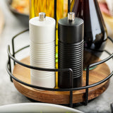 A close-up of a TableCraft Products Salt & Pepper Mill Set with white and black wood salt and pepper grinders in a metal and wooden holder, plus glass bottles of oil and vinegar, all set on a kitchen counter.