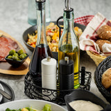 Two glass bottles of vinegar and oil, with a stylish wood salt and pepper grinder from TableCraft Products, sit in a black metal holder. Surrounding dishes include salad, bread rolls, green olives, charcuterie, and pasta on a marble table.
