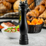 A tall TableCraft 12858 pepper mill stands on a marble countertop. Behind it, there is a white bowl of salad, a black basket filled with oranges, and another basket with assorted breads, including bagels. The background is softly blurred.