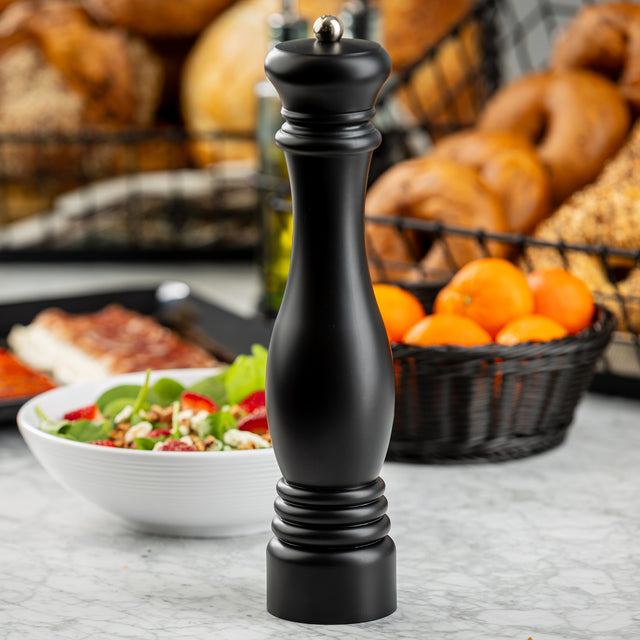 A tall TableCraft 12858 pepper mill stands on a marble countertop. Behind it, there is a white bowl of salad, a black basket filled with oranges, and another basket with assorted breads, including bagels. The background is softly blurred.