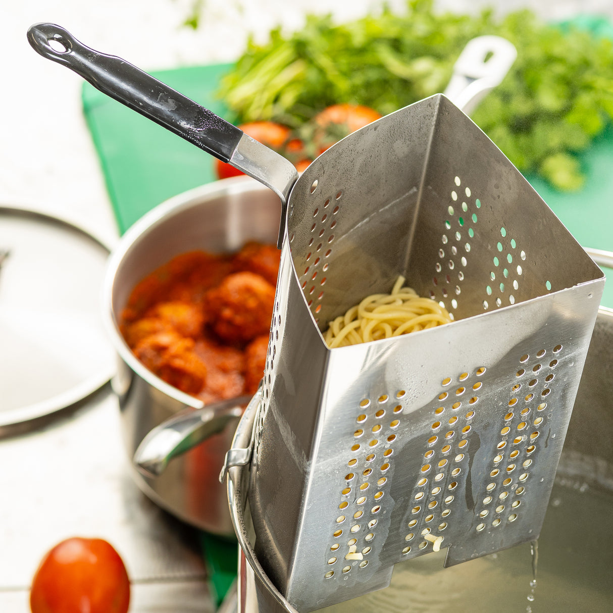 A stainless steel pasta cooker insert holds cooked spaghetti as it’s lifted from boiling water. Behind it, a pot of tomato sauce with meatballs and fresh herbs sits on a brightly lit kitchen counter alongside tomatoes and TableCraft Products.
