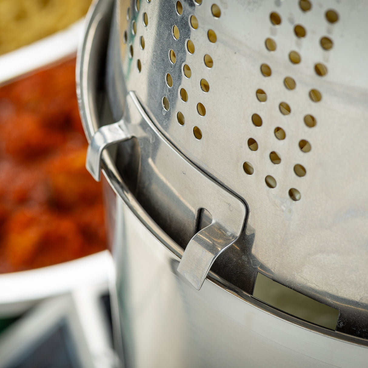Close-up of a stainless steel pasta cooker insert with small holes, locked onto a pot. In the blurred background, red pasta sauce and uncooked pasta hint at meal prep. The photo highlights the shiny metallic texture and locking mechanism.
