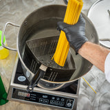 A person wearing a black glove lowers uncooked spaghetti into a large pot of boiling water using a stainless steel pasta cooker insert. The pot sits on an electric induction cooktop, with kitchen tools and TableCraft Products visible nearby.