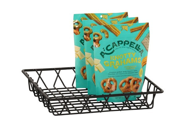A black wire basket holds three teal bags of A’Cappella Knotty Grahams, displaying pretzel-shaped graham snacks and white chocolate pieces on the packaging. The bags stand upright in the wire basket with front labels and snack images visible.