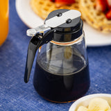 A TableCraft Modern Glass Dispenser with a black handle and metal lid sits on a blue tablecloth. In the background, waffles topped with strawberries are on a white plate, while a bowl of small round food items is in the foreground. Dishwasher safe for easy cleaning.