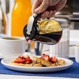 A hand pours syrup from a TableCraft Modern Glass Dispenser onto waffles topped with sliced strawberries on a white plate. A cup of coffee, a sugar container, and an orange bottle are in the background on a blue placemat.