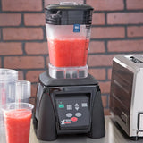 An Xtreme Series Blender with a clear pitcher blends a bright red smoothie on a gray counter. Perfect for international use, it sits beside clear cups and a stainless steel appliance, framed by a brick wall in the background.