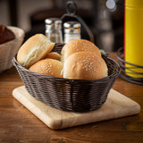 A hand-woven dark wicker basket holds four sesame seed hamburger buns, placed on a wooden cutting board. In the background are a metal condiment holder, salt and pepper shakers, and a yellow ketchup bottle on a cozy wooden table.