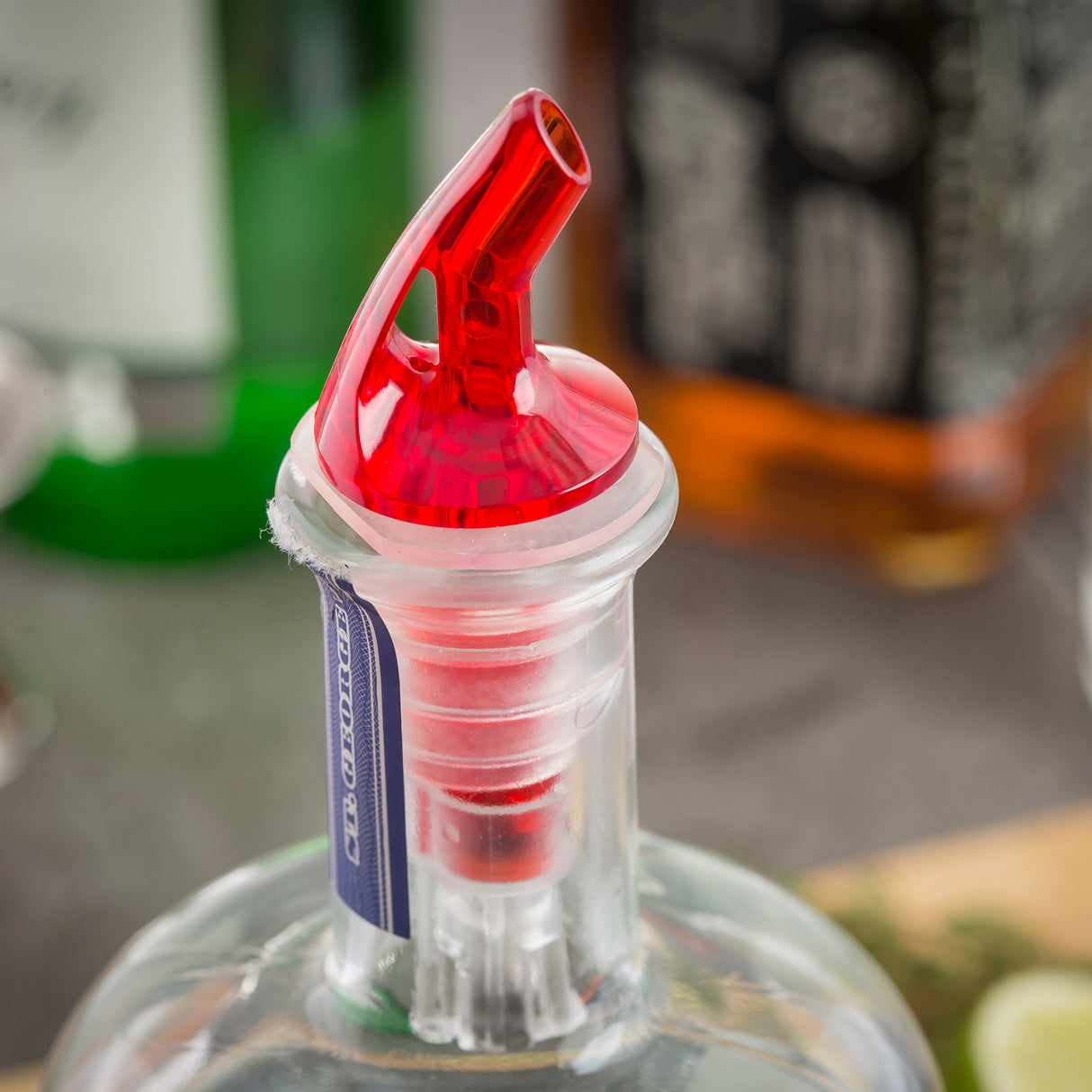 A close-up of a clear glass bottle with a red BPA free 1 oz. pourer inserted into its neck. The bottle has a partially removed blue and white tax seal, with blurred bottles and a lime wedge visible in the background.