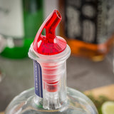 A close-up of a clear glass bottle with a red BPA free 1 oz. pourer inserted into its neck. The bottle has a partially removed blue and white tax seal, with blurred bottles and a lime wedge visible in the background.